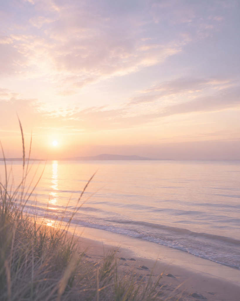 Soft sunrise over calm coastal water with gentle waves and dune grass in pastel lavender, peach, and gold tones.